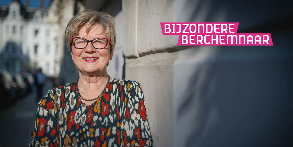 De afbeelding toont Linda die met gekruiste armen tegen een muur leunt. Ze draagt een kleurrijke top met patronen. Op de achtergrond is zijn enkele huizen uit de wijk Zurenborg zichtbaar.